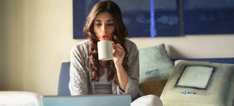 A woman with a cup of tea reading on a laptop.