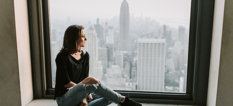 Woman in a black top and blue denim jeans sitting next to a window with the view of Empire State Building.