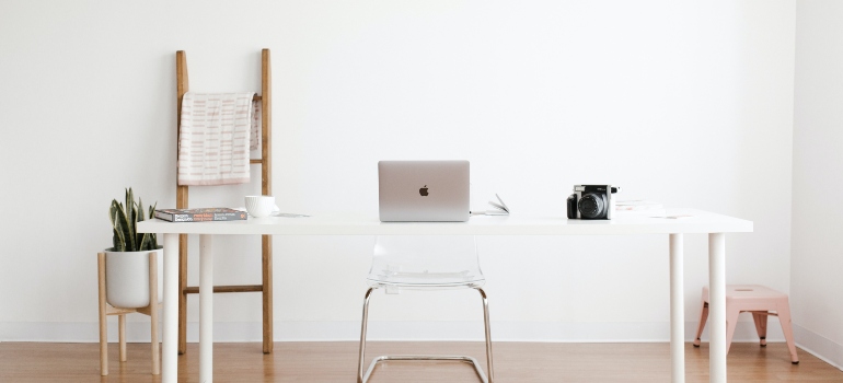 A white desk with a laptop on it in a minimalistic room with white walls.